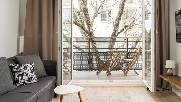A modern living room with a sofa, a small table, and a balcony with a view of trees and buildings. - Rondo Wiatraczna Harmony Apartment (Warszawa)