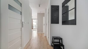 A hallway with a wooden floor, white walls, and a black bench. There are doors on the left and a large mirror on the right. - Rondo Wiatraczna Harmony Apartment (Warszawa)