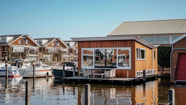 Een moderne huamboat met een dek en buitenmeubels, afgemeerd in een jachthaven met andere boten.
