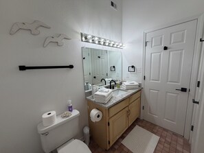 A bathroom with a toilet, sink, mirror, towel rack, and a door. - Whale Hello There . Gulf Coast Retreat . Walk to Beach . 2 Bed Duplex (Gulf Shores)