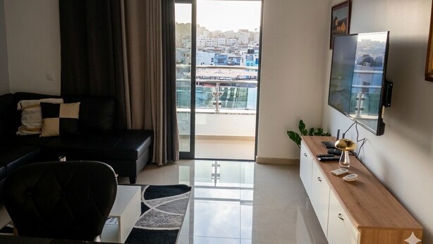 A modern living room with a sofa, a TV, a wooden cabinet, and a view of a cityscape through a large window. - Apartment Achada St Antonio Rua Principal AC, WIFI (Praia)