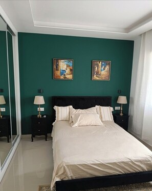 A bedroom with a bed, two paintings, two bedside tables with lamps, and a large mirror. - Apartment Achada St Antonio Rua Principal AC, WIFI (Praia)