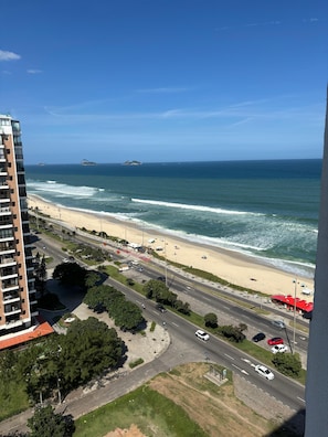 A coastal view with a beach, ocean, and a road leading to a building with multiple floors.