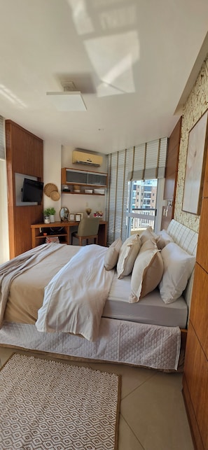 Un dormitorio con una cama, un televisor montado en la pared, un escritorio con una silla y una ventana con persianas.