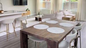 Cozinha moderna com TV de tela plana, área de jantar com mesa de madeira e jogos americanos brancos, e sala de estar com sofá e janela com cortinas.