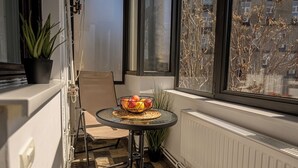 A small balcony with a table, chair, potted plant, and a window with a view of buildings.