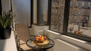 A small table with a fruit bowl, a potted plant, and a chair in a corner by a window with a view of buildings.