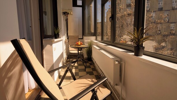 A narrow balcony with a checkered floor, a small table with a bowl of fruit, a chair, and a potted plant.