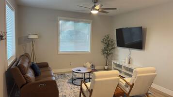 A living room with a brown leather sofa, a white armchair, a coffee table, a floor lamp, a ceiling fan, a TV, and a potted plant.