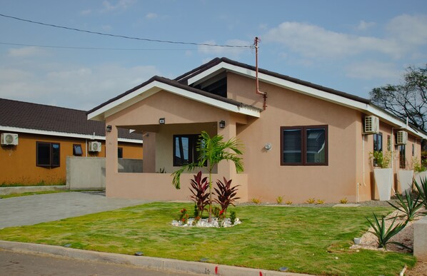 Une maison de plain-pied avec un toit plat, un petit porche à l’avant et une pelouse bien entretenue agrémentée d’un palmier et de fleurs rouges.