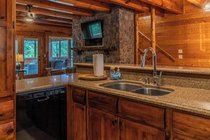 A kitchen with granite countertops, a dishwasher, a stone wall, and a television mounted above a fireplace. - 5BR/5BA Cabin | Views, Hot Tub & Pool Table (sevierville)