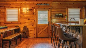 A wooden cabin interior with a dining area, bar stools, and a kitchen with a stone counter. - 5BR/5BA Cabin | Views, Hot Tub & Pool Table (sevierville)