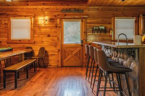 A wooden cabin interior with a dining area, bar stools, and a kitchen with a stone counter. - 5BR/5BA Cabin | Views, Hot Tub & Pool Table (sevierville)
