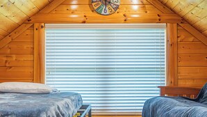 A cozy attic bedroom with wooden walls, a sloped ceiling, a bed with a headboard, a bench, a clock, and a window with blinds. - 5BR/5BA Cabin | Views, Hot Tub & Pool Table (sevierville)
