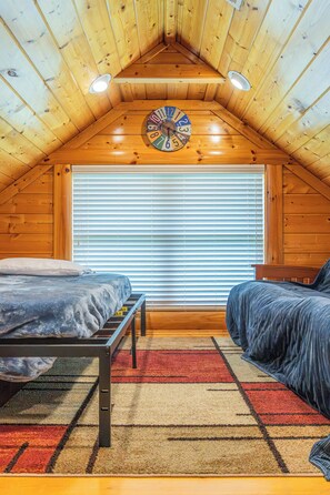 A cozy attic bedroom with wooden walls, a sloped ceiling, a bed with a headboard, a bench, a clock, and a window with blinds. - 5BR/5BA Cabin | Views, Hot Tub & Pool Table (sevierville)