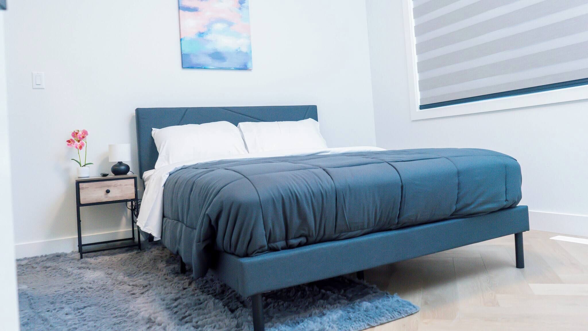 A bedroom with a bed, a nightstand, a lamp, a painting, and a window with blinds.