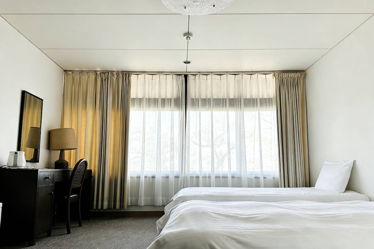 Une chambre d’hôtel avec un lit, un bureau, une chaise, une lampe et une fenêtre avec des rideaux.
