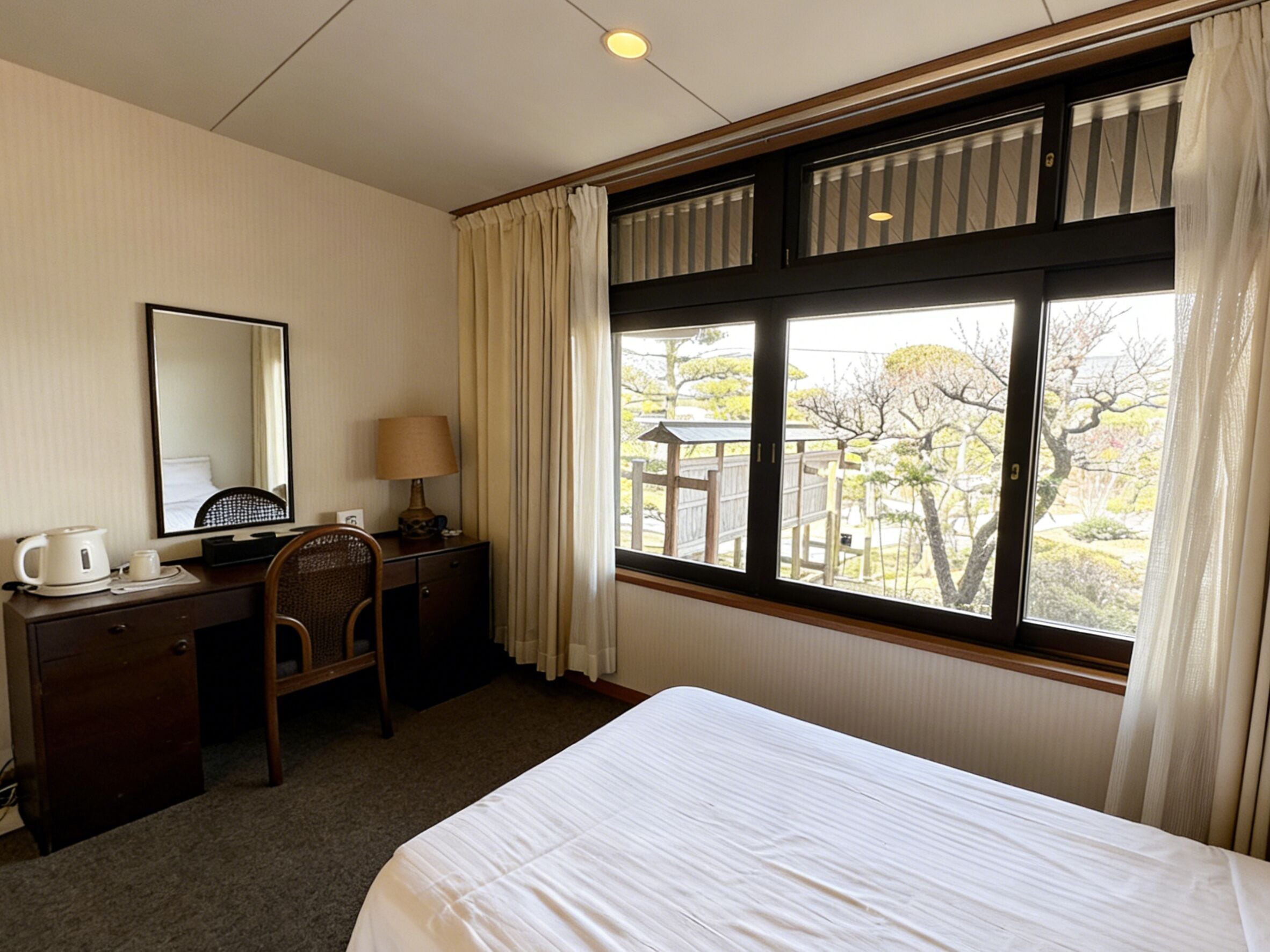 Habitación de hotel con cama, escritorio con silla, lámpara, cafetera y ventana con cortinas.