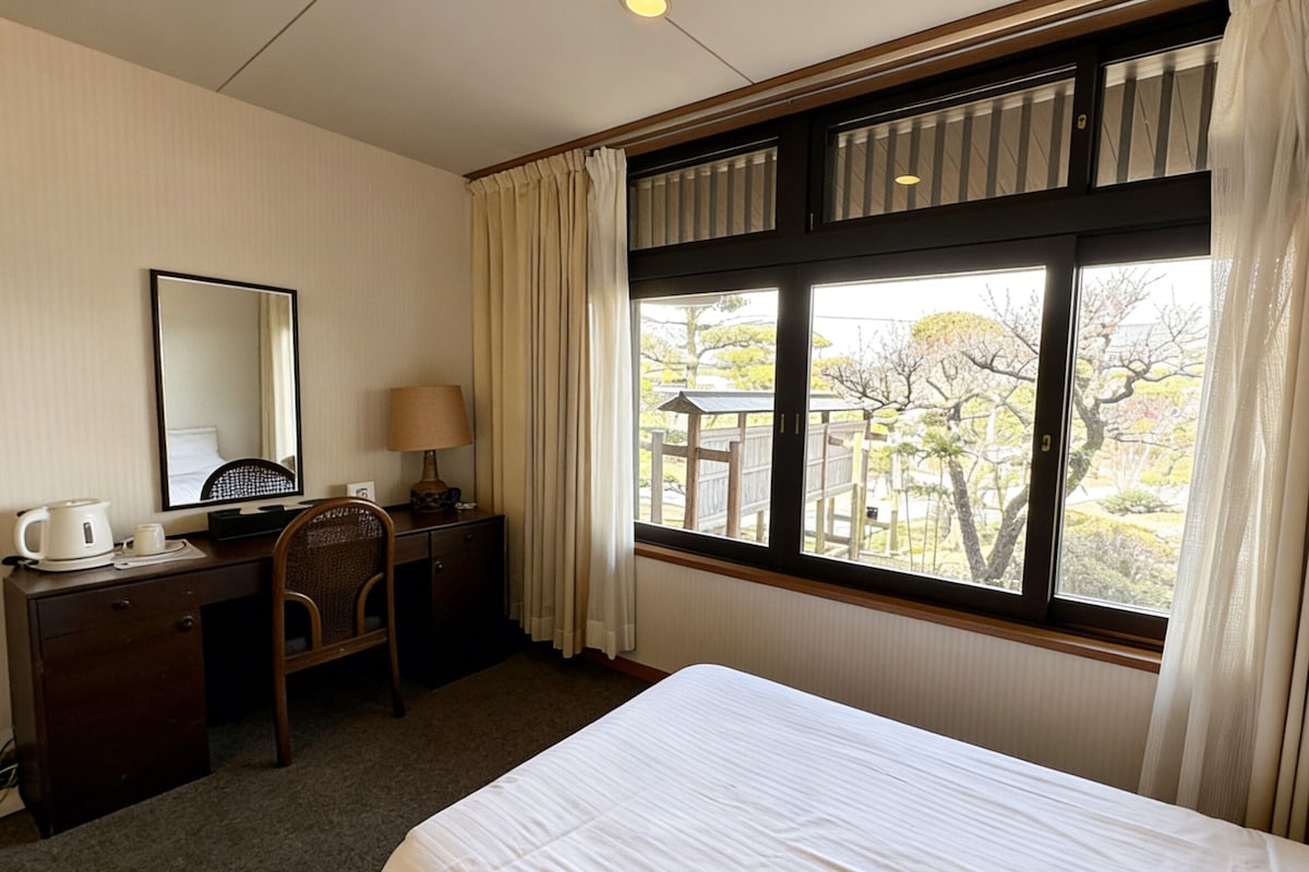 A hotel room with a bed, a desk with a chair, a lamp, a coffee maker, and a window with curtains.