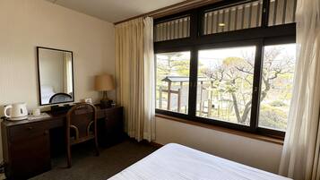 Habitación de hotel con cama, escritorio con silla, lámpara, cafetera y ventana con cortinas.