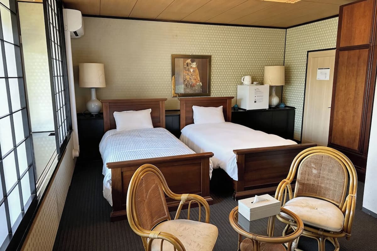 Une chambre d’hôtel avec deux lits, un bureau, deux chaises, une petite table, un tableau au mur et une fenêtre.