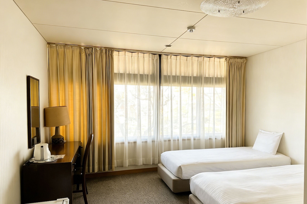 A hotel room with two beds, a desk with a lamp, a chair, and a large window with curtains.