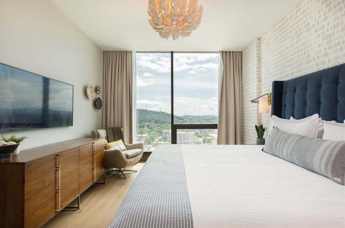 Une chambre d’hôtel moderne avec un grand lit, un téléviseur à écran plat, une commode en bois, une chaise et une vue sur la ville.