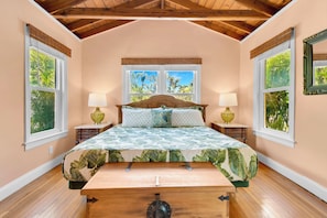 Ein Schlafzimmer mit Holzboden, einem Bett mit blattgemusterten Bettwäsche, zwei Nachttischlampen und Fenstern mit Blick auf Grünflächen.