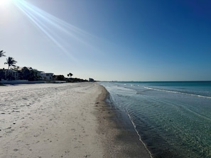 Ein Strand mit klarem Wasser, sandigem Ufer und Gebäuden im Hintergrund.