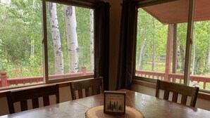 A wooden dining table with a framed picture, surrounded by chairs, in a room with large windows overlooking a forest. - Two Houses, Twice the Fun (Glacier County)