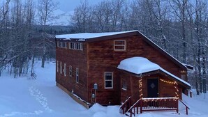 A wooden cabin with a snow-covered roof, surrounded by trees, and a porch with lights. - Two Houses, Twice the Fun (Glacier County)