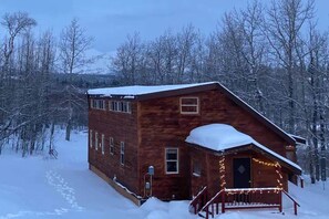 A wooden cabin with a snow-covered roof, surrounded by trees, and a porch with lights. - Two Houses, Twice the Fun (Glacier County)