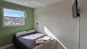 Ein Schlafzimmer mit Bett, Fenster, an der Wand montiertem Fernseher und Blick auf Gebäude.