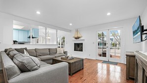 Amplia sala de estar con sofá seccional gris, una mesa de centro de madera y una chimenea. Hay un ventanal con vista a la playa y una puerta que da a un balcón.