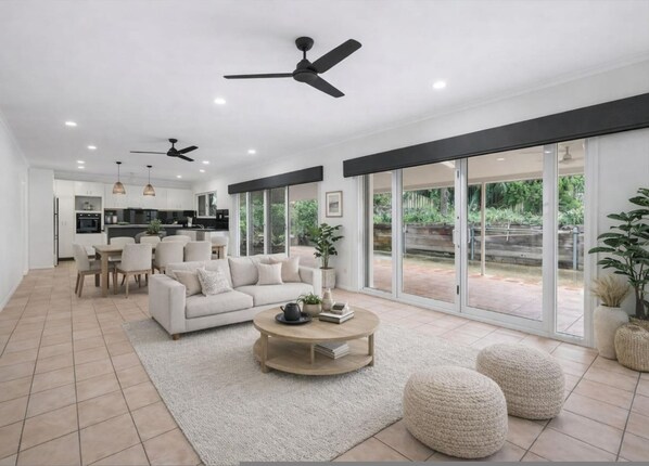 A modern living room with a sofa, coffee table, and ottomans, a kitchen in the background, and large sliding glass doors leading to a garden. - Cullen Bay Retreat Pool Balcony Marina Stroll (Darwin)