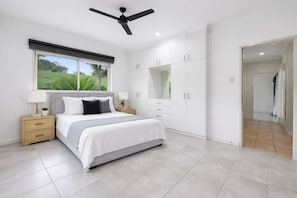 Une chambre à coucher comprenant un lit, des tables de chevet, un ventilateur de plafond, une grande armoire et une fenêtre avec des stores.
