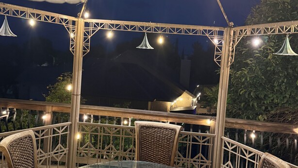 An outdoor patio with a glass table surrounded by wicker chairs, string lights, and a metal railing. - Home With A View, Beautiful 3 Bedroom 4 Bath, Each Bedroom with Private Bath (Redding)