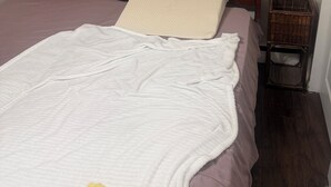 Un lit avec une tête de lit en bois, une couverture blanche, une serviette jaune et une table de chevet en bois avec un panier.