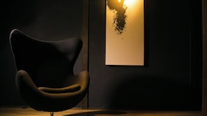 A modern chair with a curved design and a metallic base, positioned on a wooden floor with a dark wall in the background.