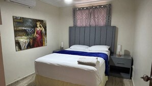 Un dormitorio con una cama, una mesita de noche, aire acondicionado instalado en la pared, un cuadro y una cortina estampada.
