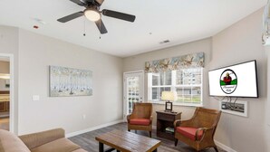 Ein Wohnzimmer mit einer Couch, zwei Korbstühlen, einem Couchtisch, einem Deckenventilator, einem Gemälde, einem Fenster mit Jalousien und einem an der Wand montierten Fernseher.
