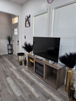Una sala de estar con un televisor de pantalla plana grande sobre un soporte de madera, una puerta con ventana y una planta en maceta.