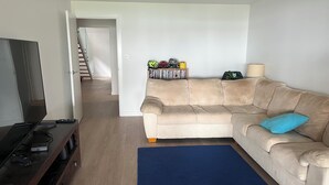 Ein Wohnzimmer mit Fernseher, einem beigen Sofa, einem blauen Teppich und einem Bücherregal mit diversen Gegenständen.