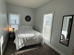 A bedroom with a bed, a nightstand with a lamp, a wall clock, and a window with blinds.