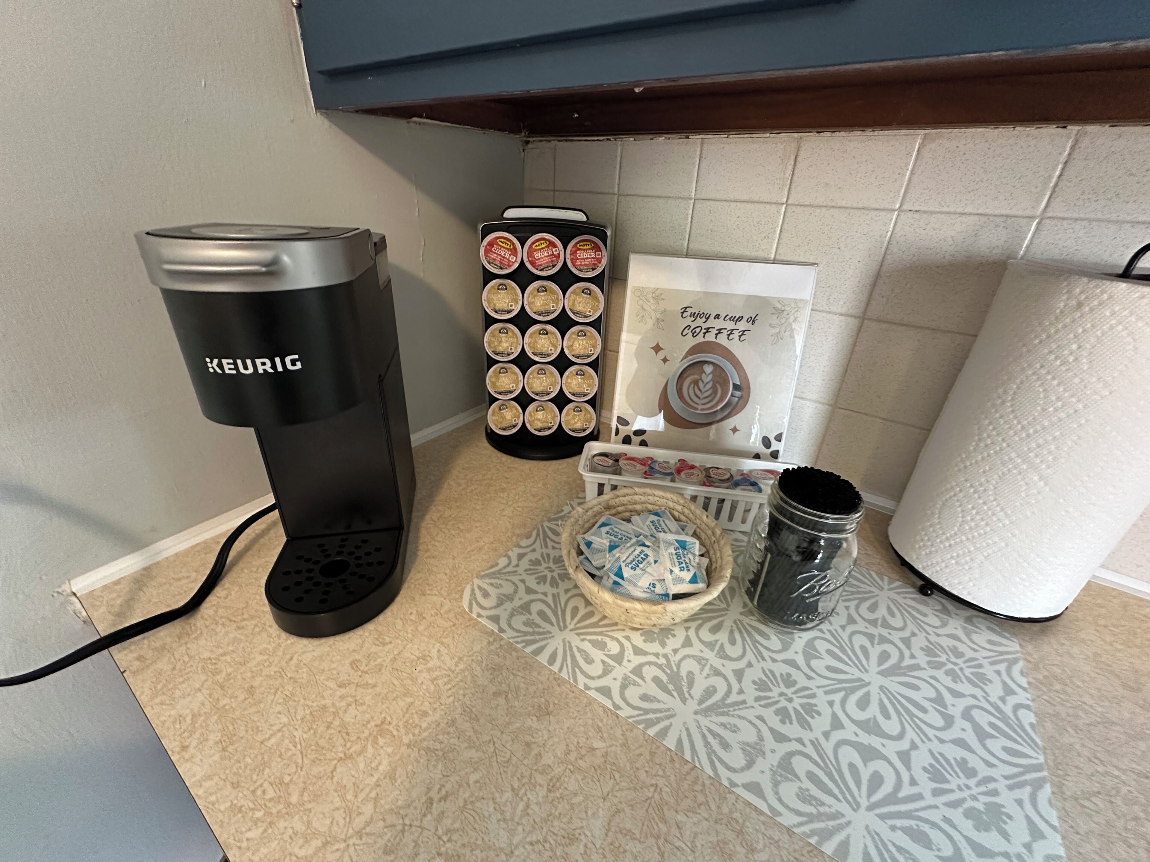 A kitchen counter with a Keurig coffee maker, a coffee pod holder, a basket of sugar packets, a jar of coffee grounds, and a roll of paper towels.