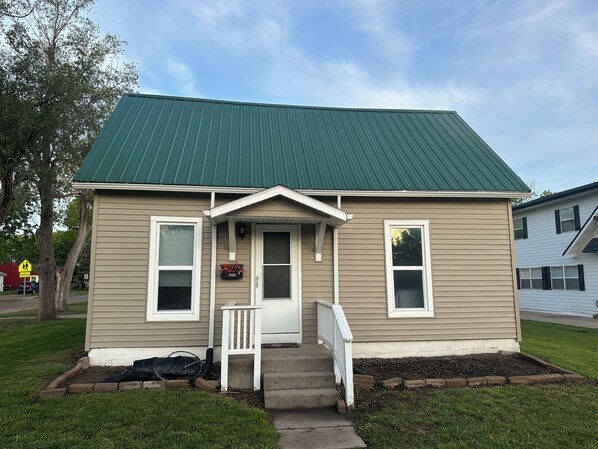A single-story house with a green metal roof, a front porch with a white railing, and a small front yard.