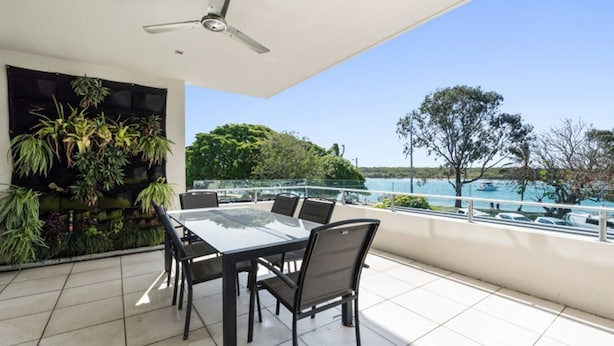 Un balcon avec une table et des chaises, un ventilateur de plafond et une vue sur un lac où naviguent des bateaux.