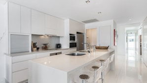 Une cuisine moderne avec des meubles blancs, un grand îlot central doté de tabourets, des appareils en acier inoxydable et une vue sur le salon attenant.