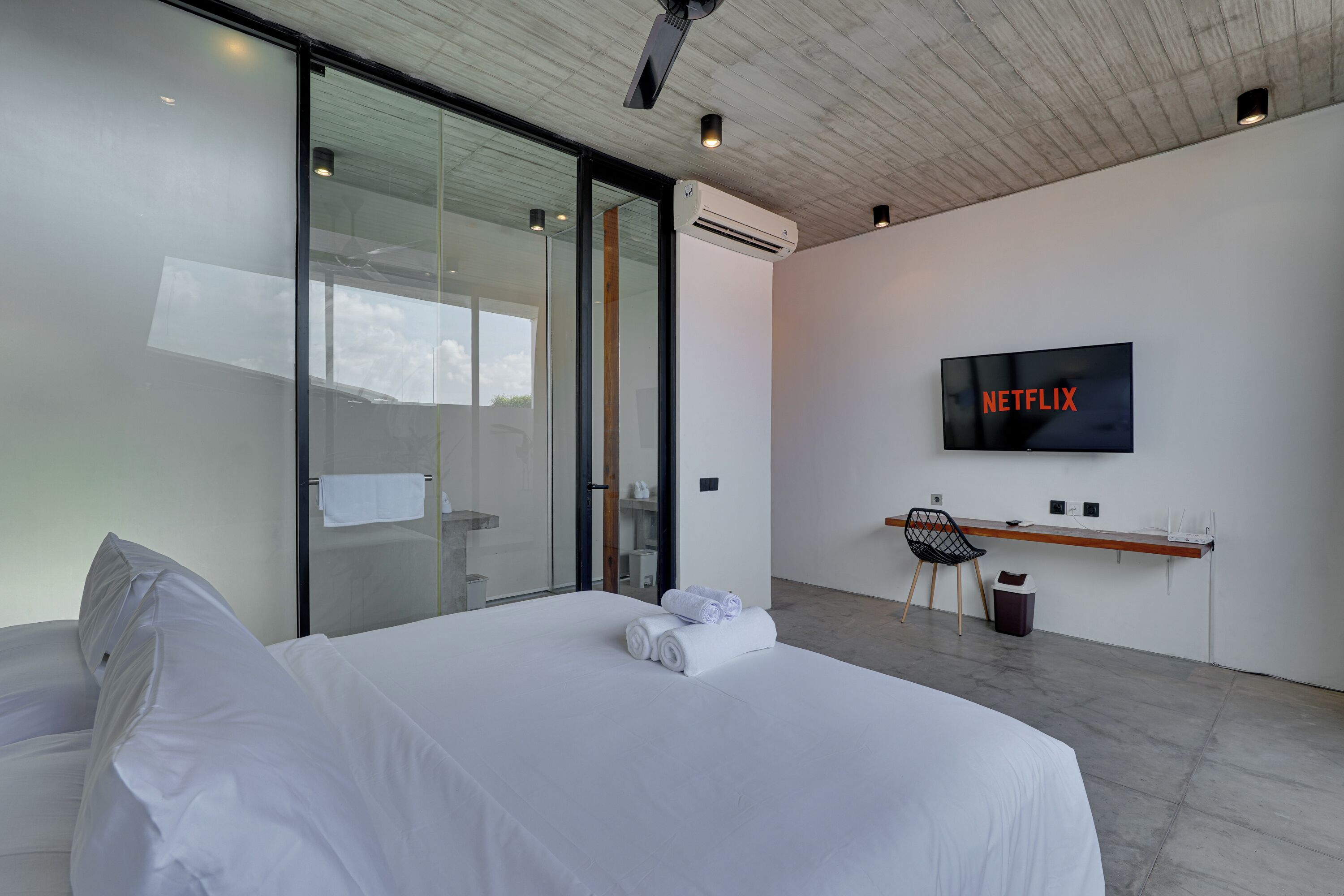 A modern hotel room with a bed, a desk, a chair, a TV, and an air conditioning unit.