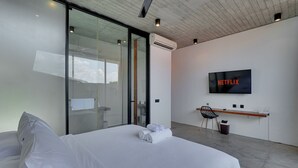 A modern hotel room with a bed, a desk, a chair, a TV, and an air conditioning unit. - Rhys Apartment - Berawa (Canggu)